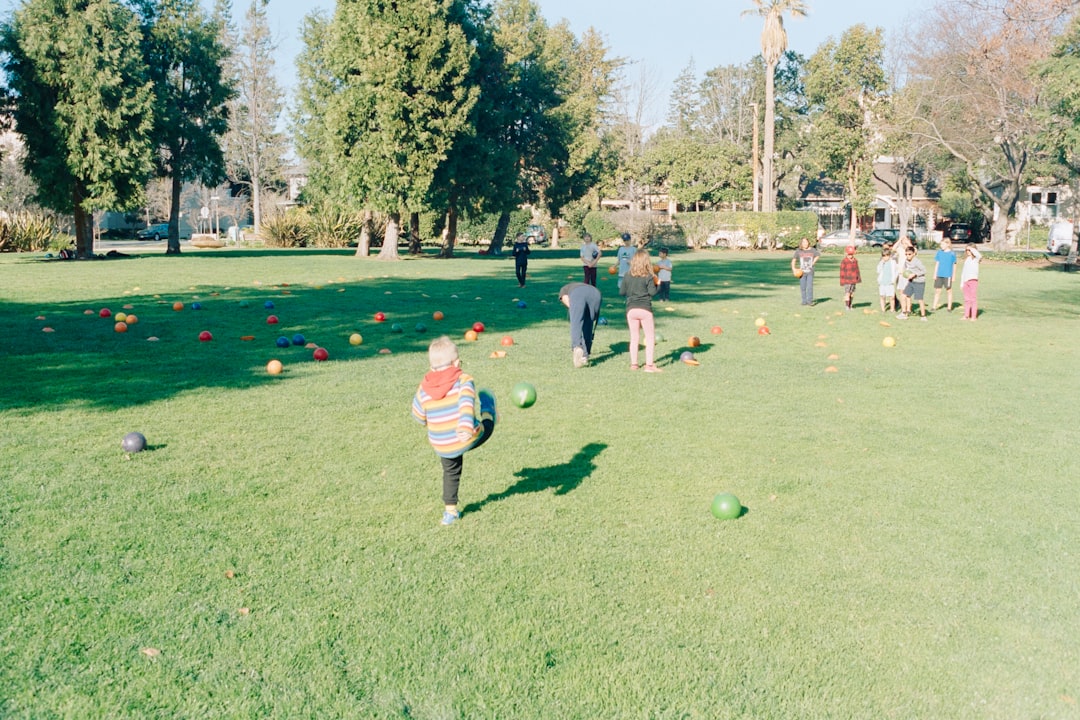 A group of children and adults playing with colorful balls and cones on a sunny green lawn in a park with large trees in the background